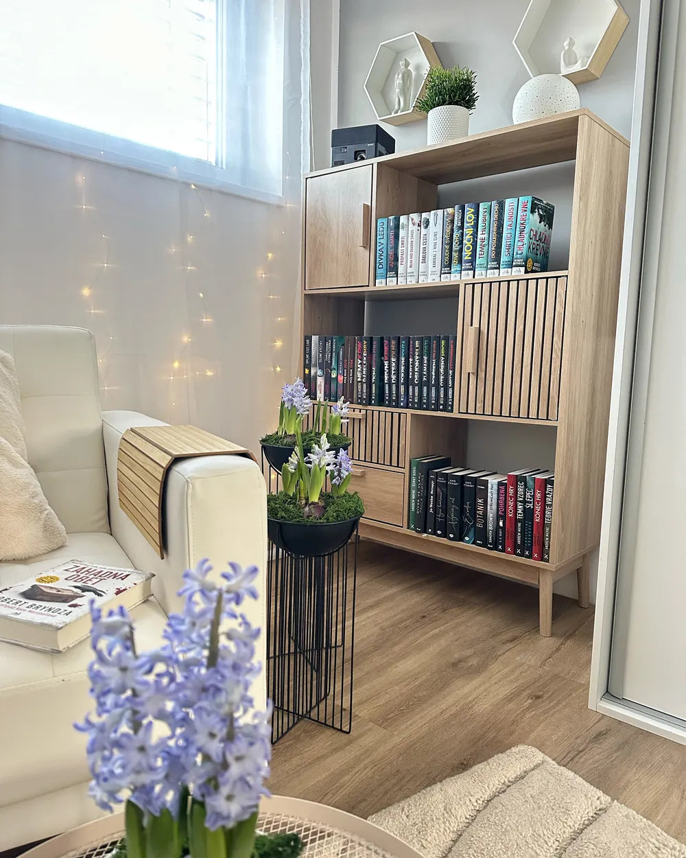 A light wood bookcase with slatted doors and open shelves, styled with books and small plants in a bright living room corner next to a white sofa.