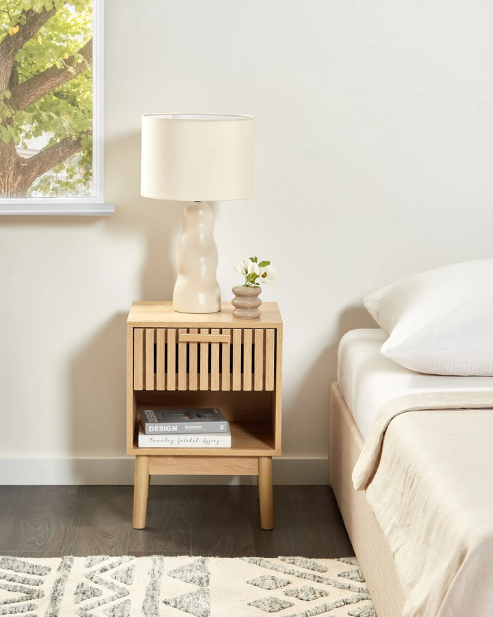 Wooden nightstand with lamp and books next to a bed in a bedroom.