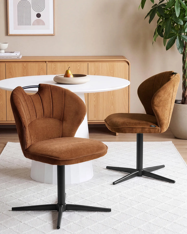 Two modern swivel dining chairs upholstered in warm brown velvet, shown in a styled dining room setting with a white table, light wood cabinet, and a potted plant.