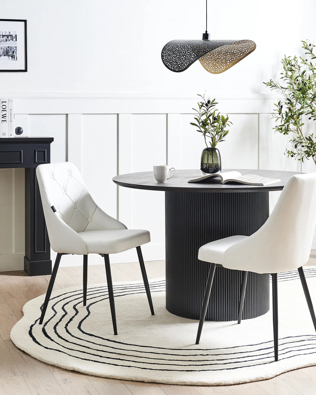 A modern dining room scene featuring a round black ribbed pedestal table with two white upholstered chairs, set on a cream rug with black wavy lines. A black pendant light hangs above, and a fireplace and greenery decor are visible in the background.