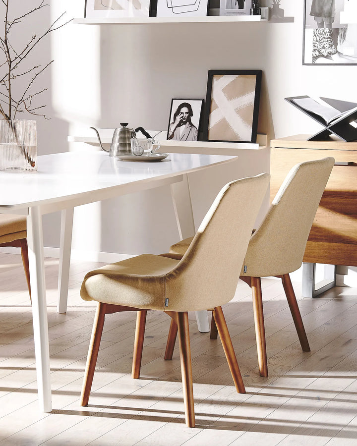 Two modern dining chairs with beige upholstered seats and light brown wooden legs, positioned in a bright, contemporary dining room next to a white table.
