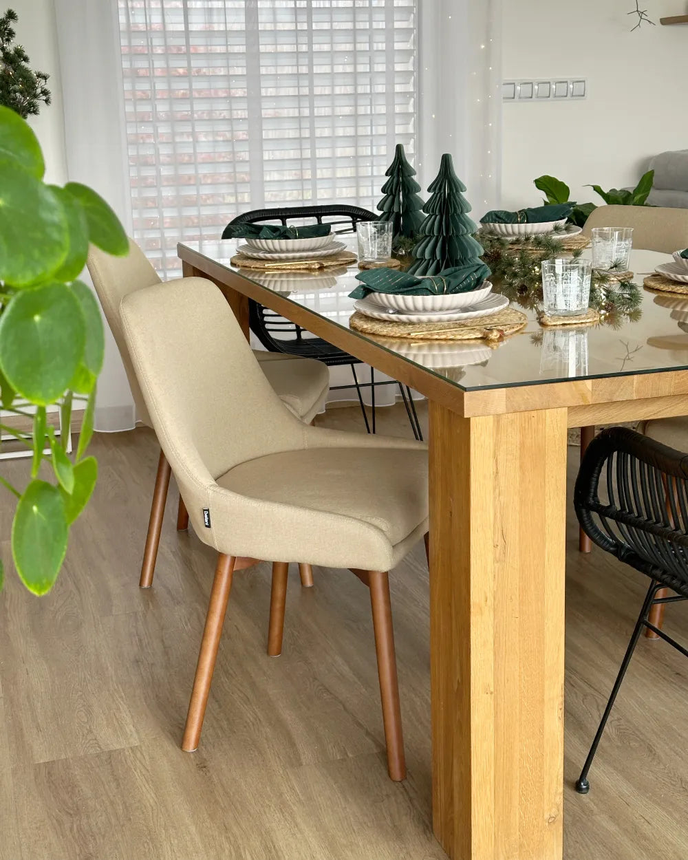 A modern dining room scene featuring a light oak wood dining table with a clear glass top, surrounded by beige upholstered dining chairs with light wood legs, set on a light wood floor.