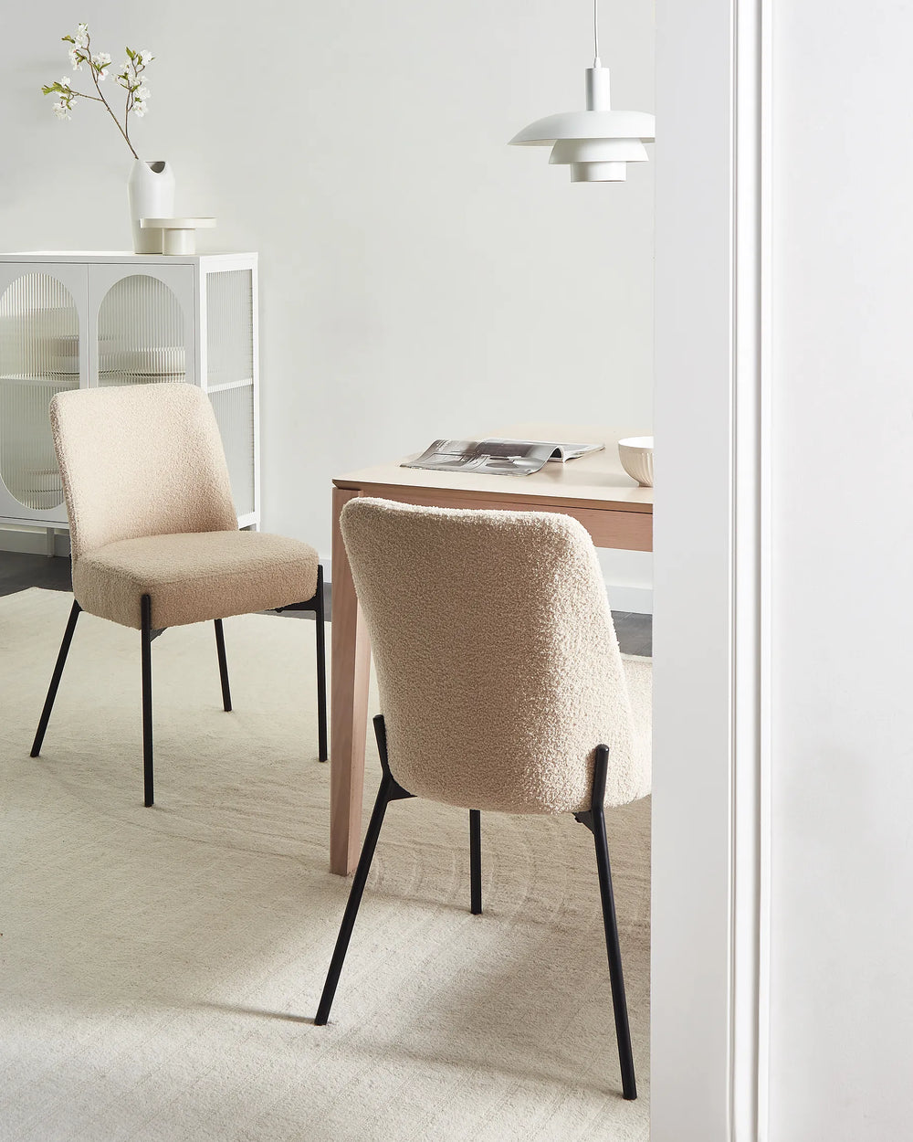 Two upholstered dining chairs in a warm beige bouclé fabric with black metal legs, shown in a bright, modern dining room setting next to a wooden table and pendant light.