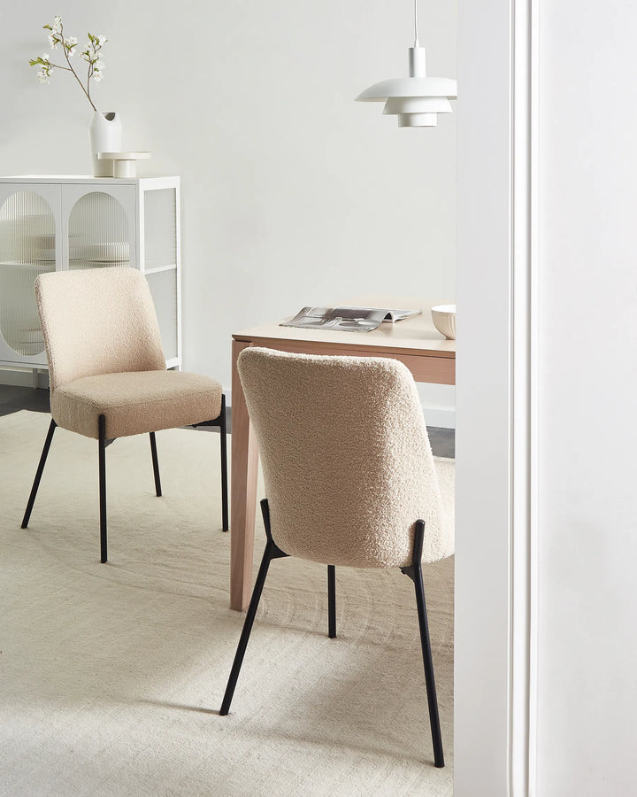 Two upholstered dining chairs in a warm beige bouclé fabric with black metal legs, shown in a bright, modern dining room setting next to a wooden table and pendant light.