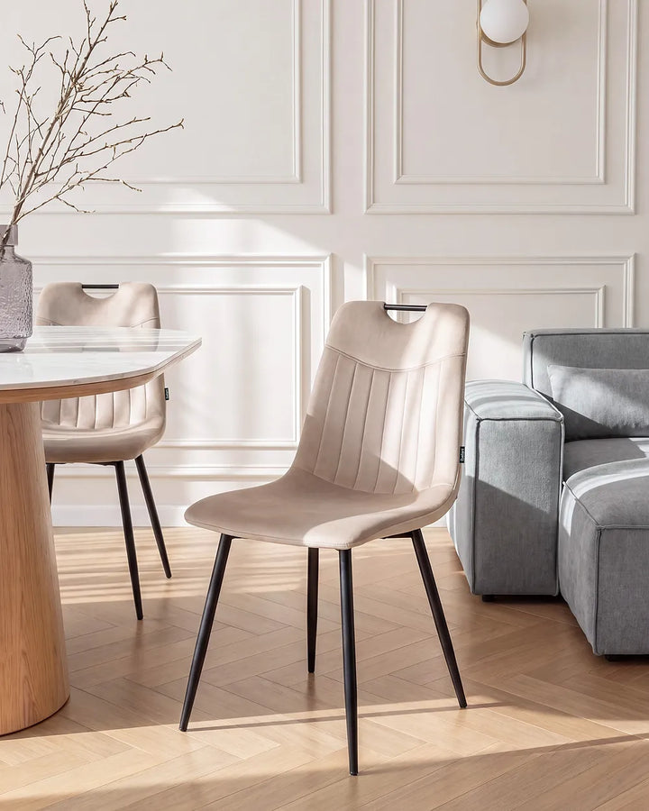 A modern dining chair upholstered in a light beige fabric with black tapered legs, shown in a bright, contemporary dining room setting next to a gray sofa and wooden table.