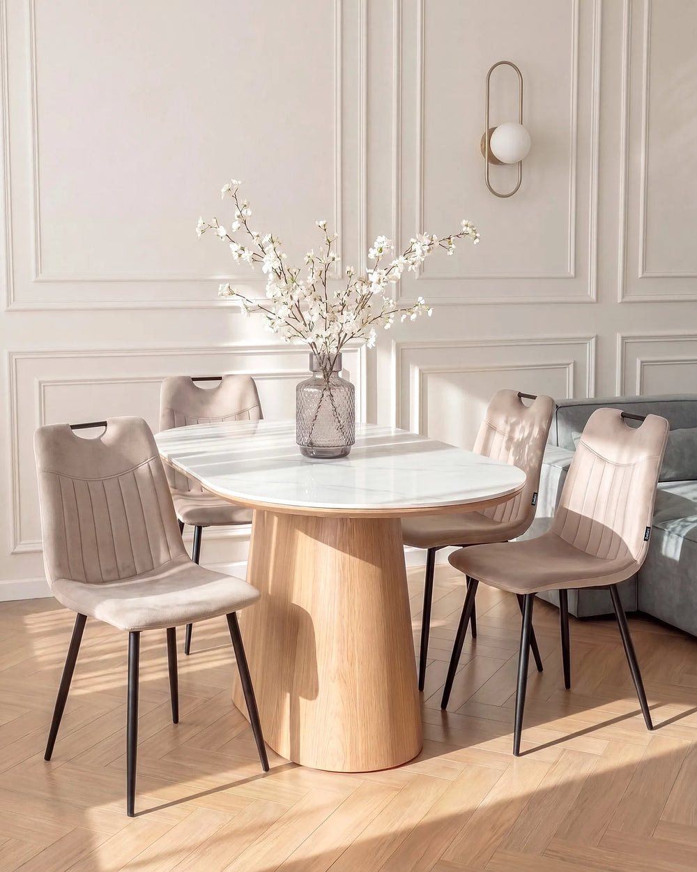 A round dining table with a white marble top and a natural wood pedestal base, surrounded by four blush pink upholstered dining chairs with black metal legs. The set is styled in a bright, modern dining room with cream paneled walls, a gold wall sconce, and light wood herringbone flooring.
