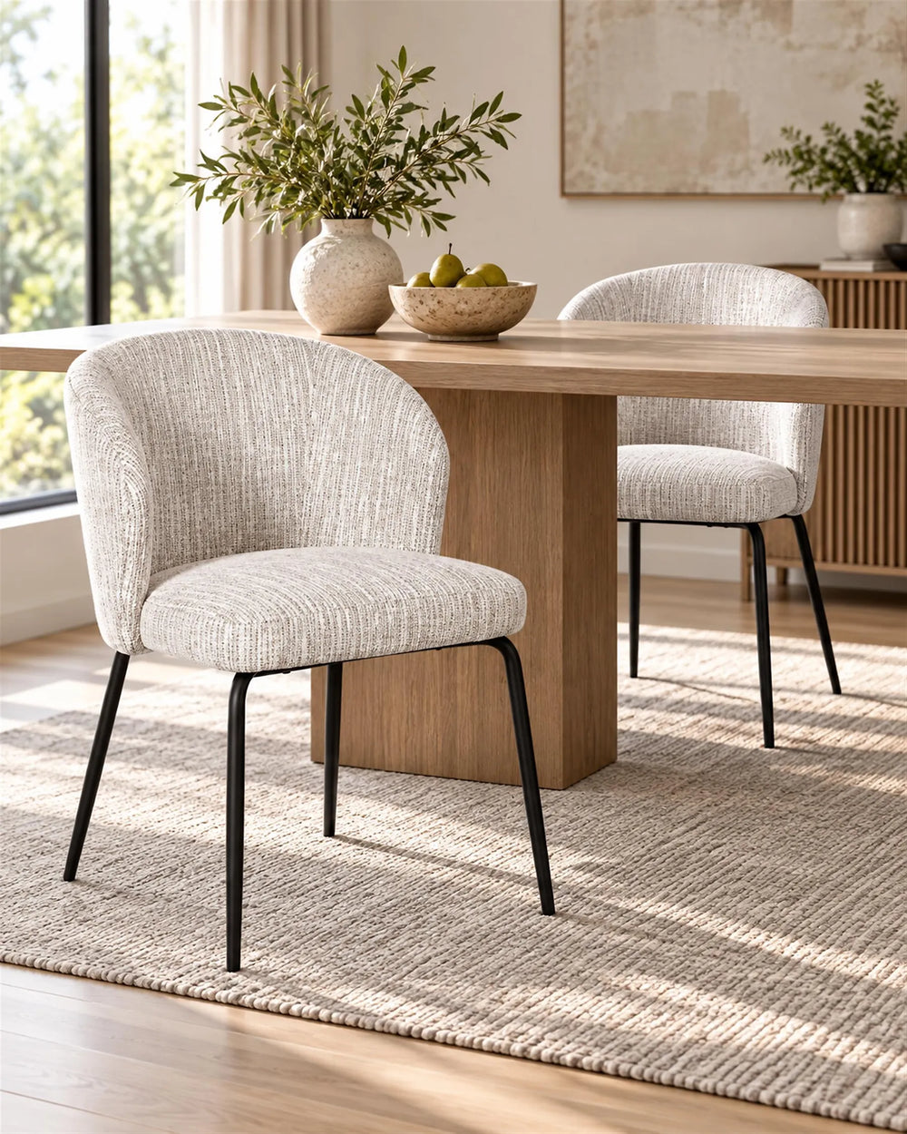 Two upholstered dining chairs with textured light beige fabric and black metal legs, shown around a wooden dining table in a bright, modern dining room.