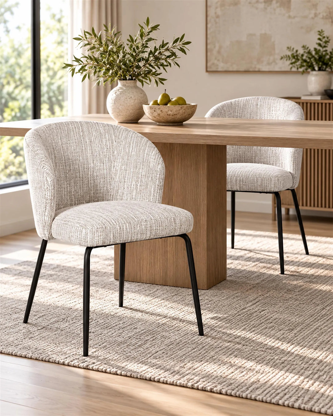 Two upholstered dining chairs with textured light beige fabric and black metal legs, shown around a wooden dining table in a bright, modern dining room.