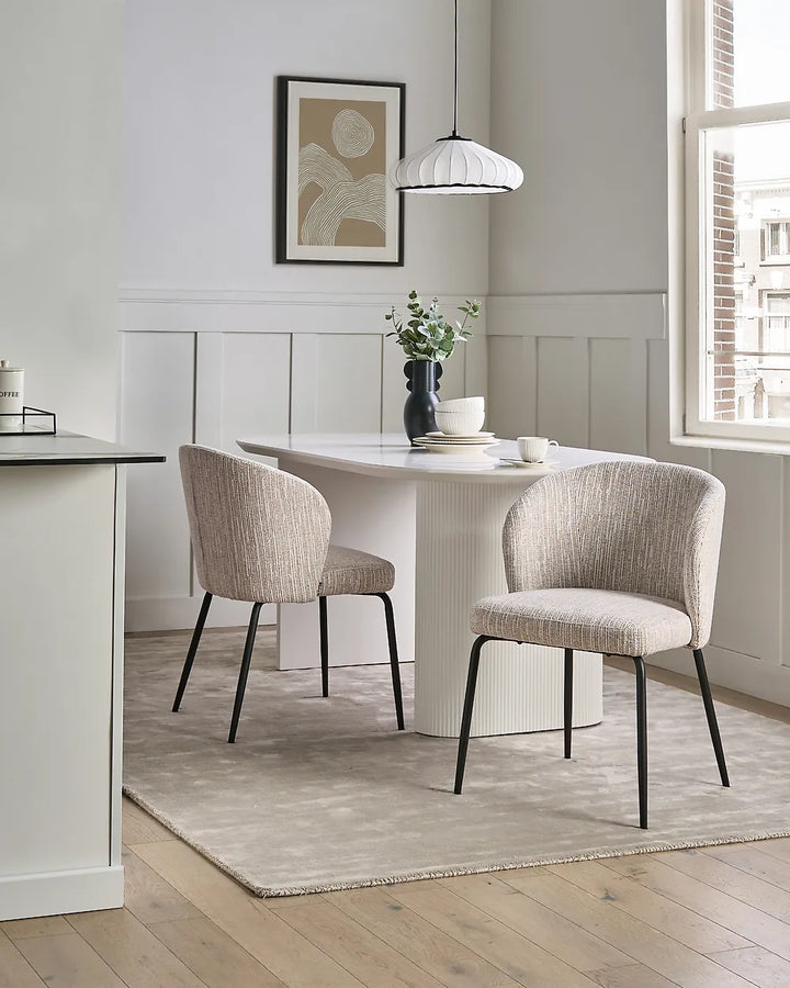 A dining area featuring two upholstered accent chairs with black metal legs around a white pedestal table on a light beige rug. The room has white paneled walls, light wood floors, a hanging pendant light, and a framed abstract print.