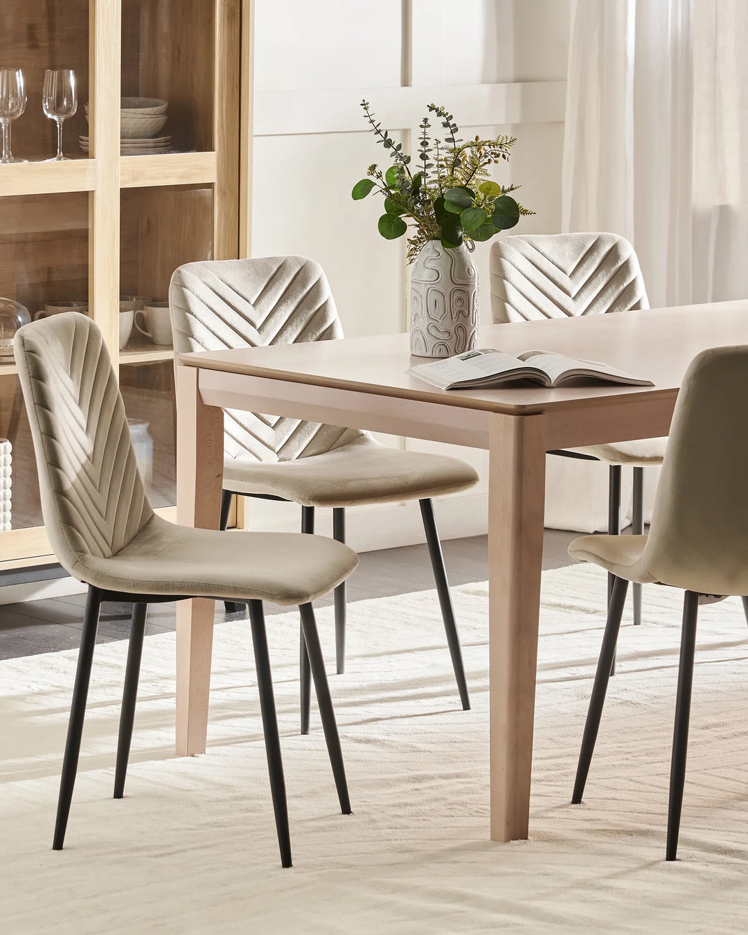 A light wood dining table with four upholstered chairs in a neutral beige and cream color palette, styled with a vase of greenery and an open book on a light-colored rug.