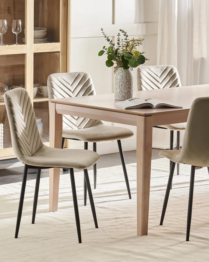 A light wood dining table with four upholstered chairs in a neutral beige and cream color palette, styled with a vase of greenery and an open book on a light-colored rug.
