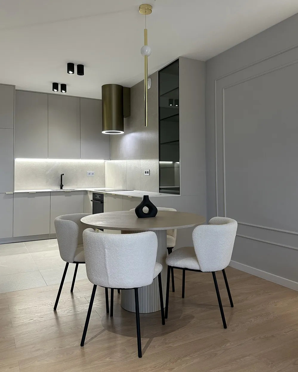 A modern dining area featuring a round light wood table with four white upholstered chairs, set in a contemporary kitchen with gray cabinets, a gold range hood, and light wood flooring.