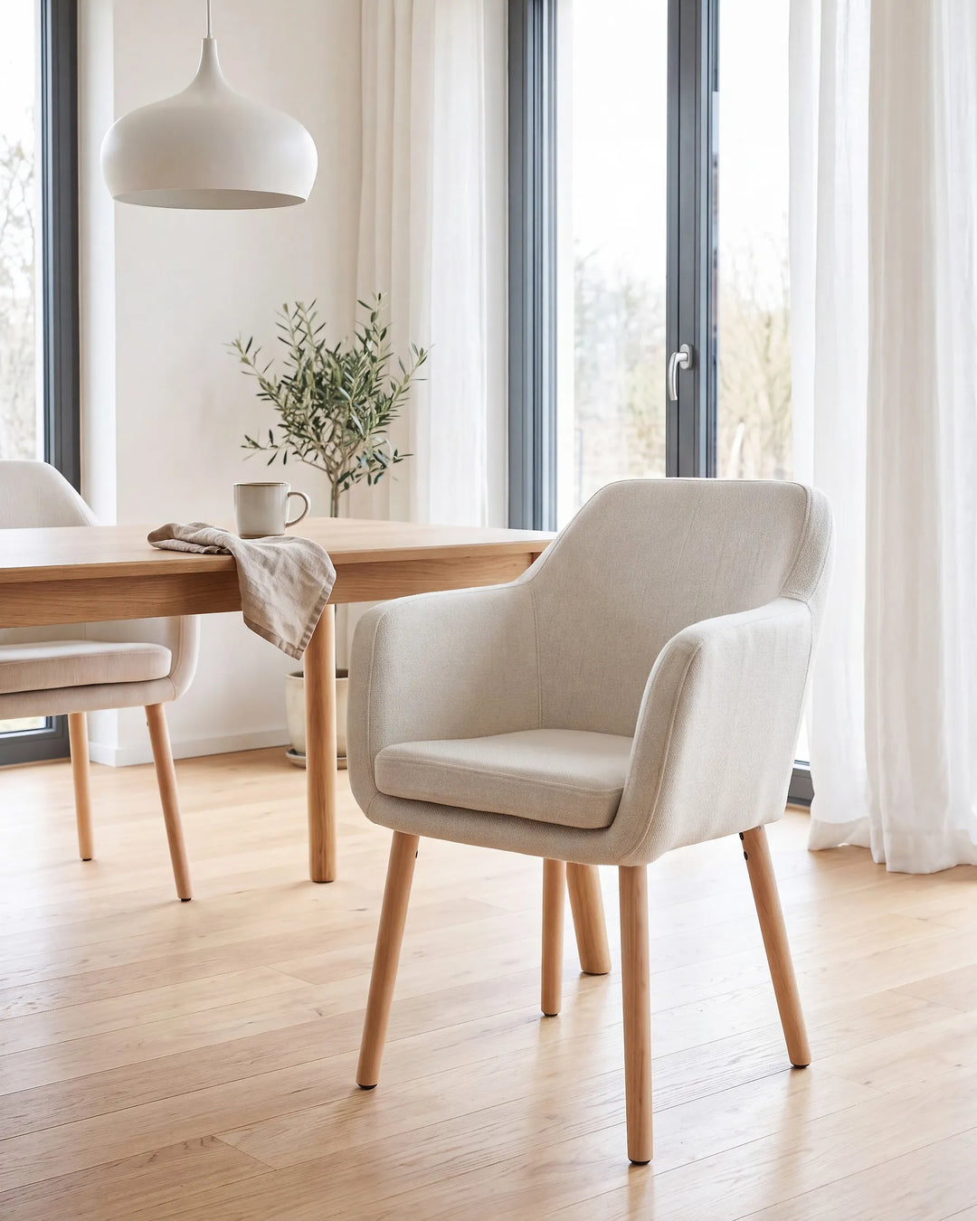 A modern upholstered dining chair in a light beige fabric with light wood legs, shown in a bright, contemporary dining room next to a wooden table.