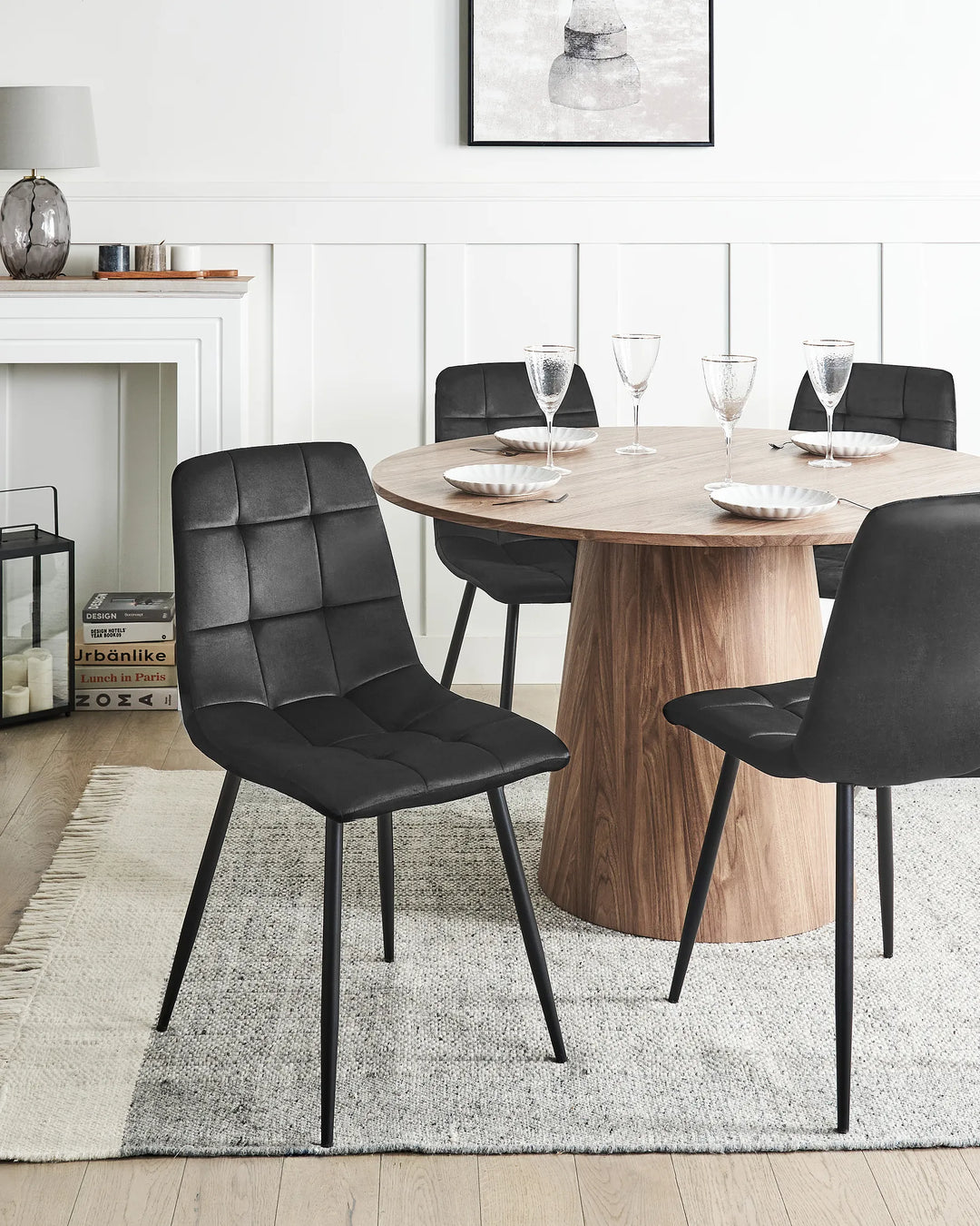 A modern dining room scene featuring a round wooden table surrounded by four black upholstered dining chairs with thin black metal legs, set on a light gray rug.