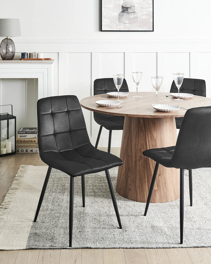 A modern dining room scene featuring a round wooden table surrounded by four black upholstered dining chairs with thin black metal legs, set on a light gray rug.