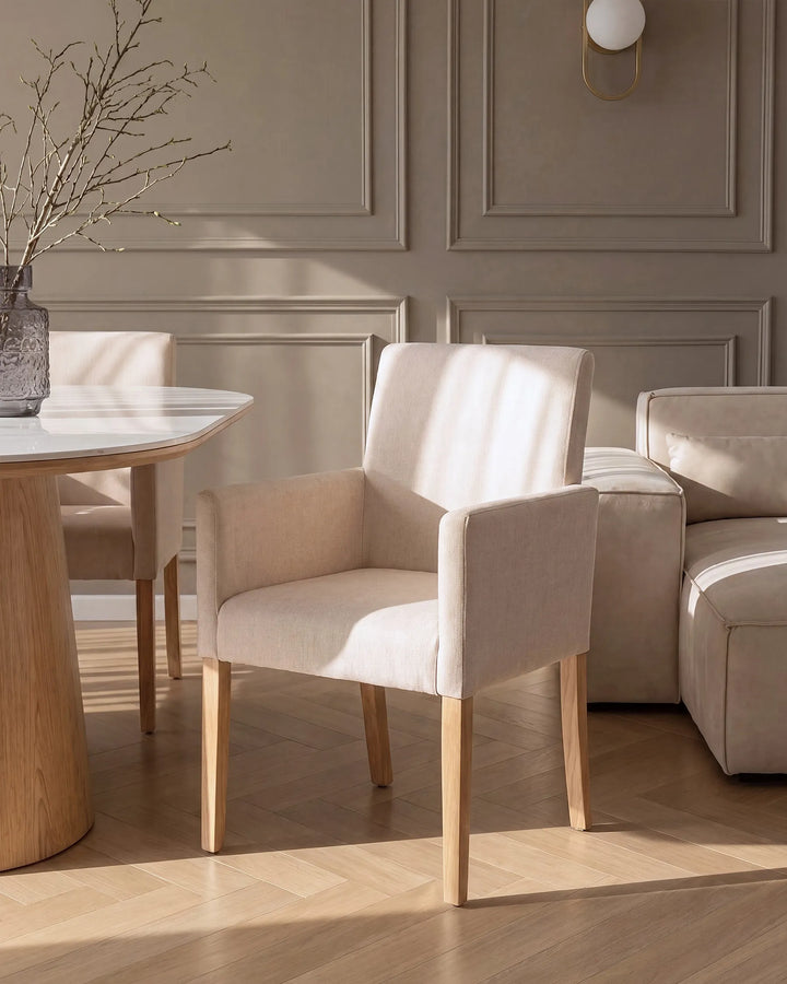 A light beige upholstered dining chair with light wood legs, shown in a styled dining room setting with a marble-top table, matching chair, sofa, and paneled wall.