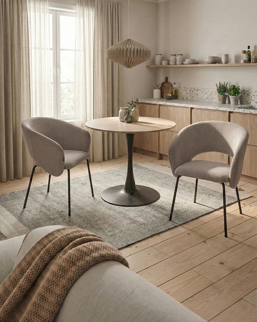 A modern dining set featuring a round light wood table with a black pedestal base, flanked by two gray upholstered accent chairs on a textured gray rug in a bright, contemporary dining room.