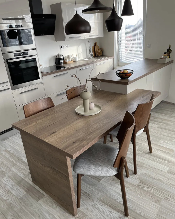 A modern kitchen island with a rectangular wood-look dining table and four wooden chairs with gray upholstered seats, set in a bright contemporary kitchen with light wood floors.