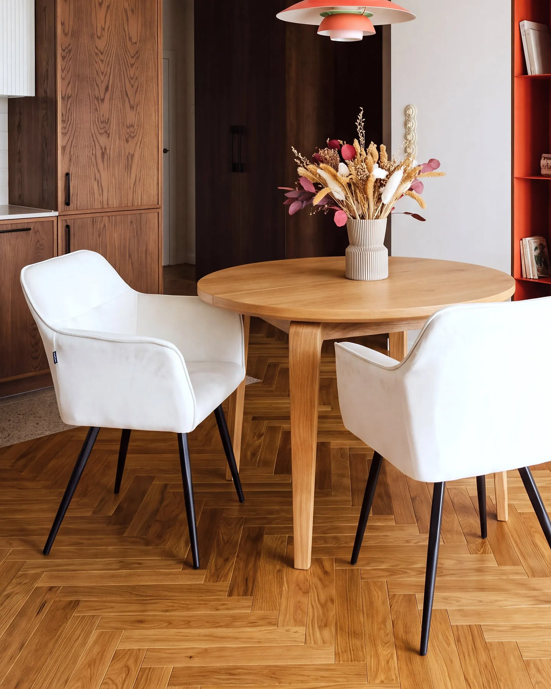 A round light oak dining table with two white upholstered chairs and black legs, styled in a modern dining room with a wooden herringbone floor and a vase of dried flowers on top.