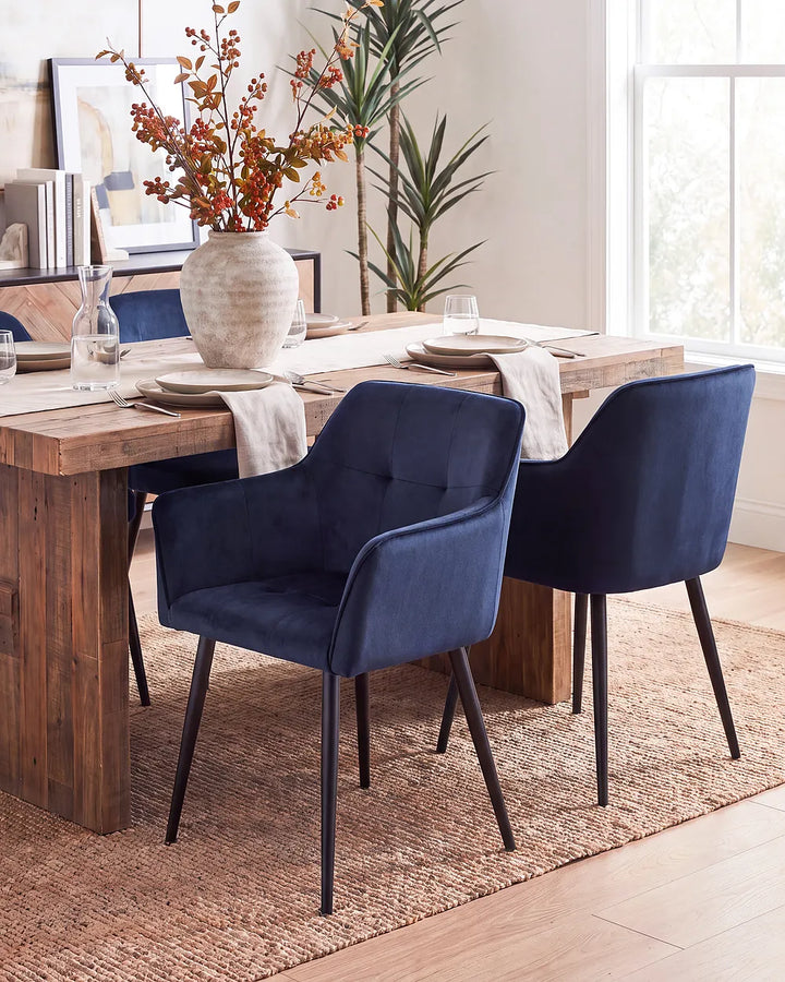 Two navy blue upholstered dining chairs with black metal legs, shown around a rustic wooden dining table in a bright, modern dining room setting.