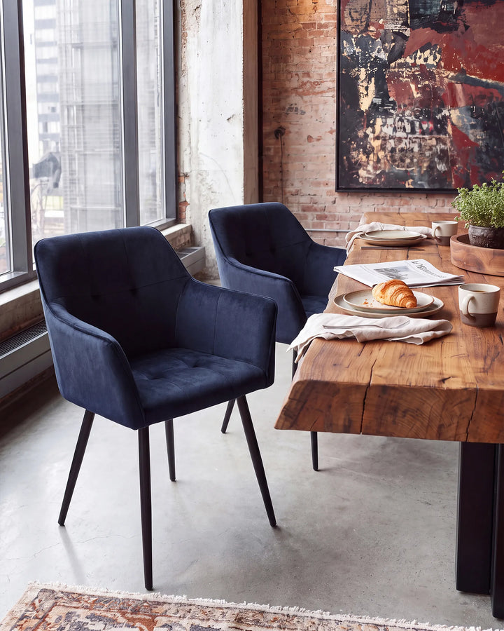 Two modern navy blue velvet dining chairs with black metal legs, positioned in a stylishly decorated loft dining room next to a rustic wooden table.