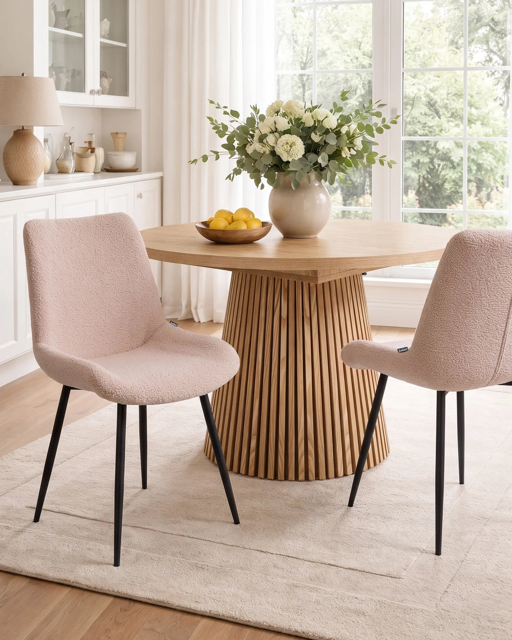 A dining room scene with a round light wood table featuring a slatted pedestal base, two blush pink upholstered chairs with black legs, and a large vase of white flowers on the table.