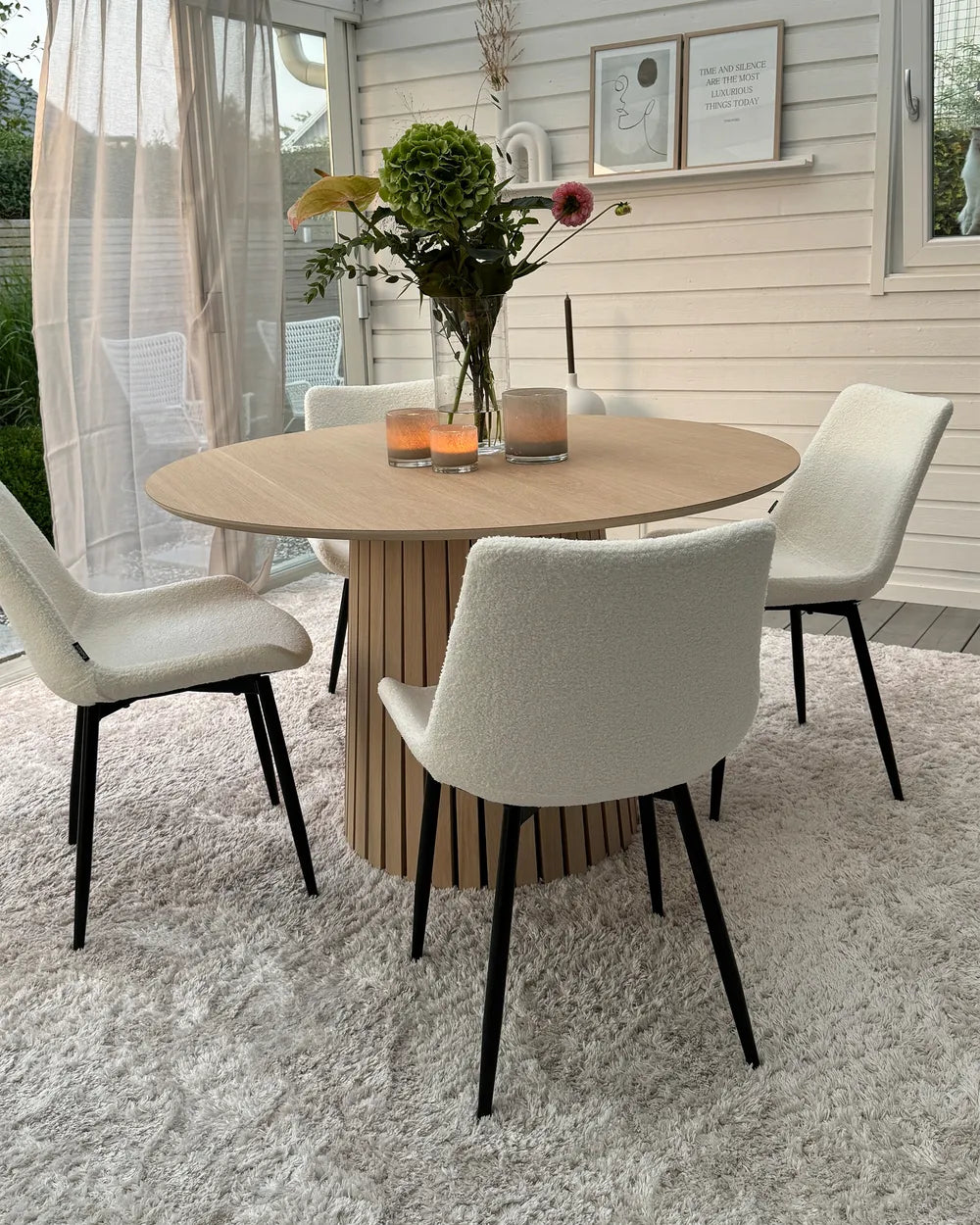 A round dining table with a light wood top and slatted wood pedestal base, surrounded by four upholstered dining chairs with cream bouclé seats and black legs, set on a white shag rug in a bright dining room.