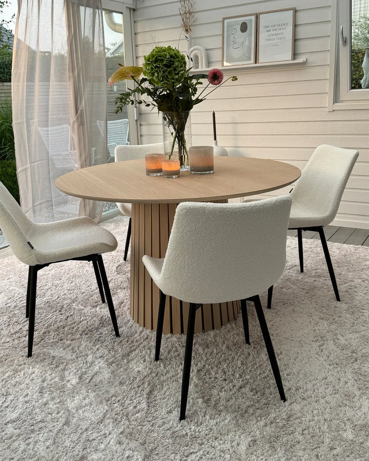 A round dining table with a light wood top and slatted wood pedestal base, surrounded by four upholstered dining chairs with cream bouclé seats and black legs, set on a white shag rug in a bright dining room.