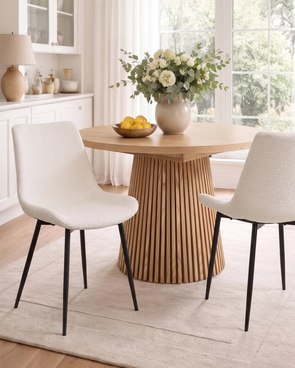 A round wooden dining table with a fluted pedestal base, styled with two white upholstered chairs in a bright, modern dining room.