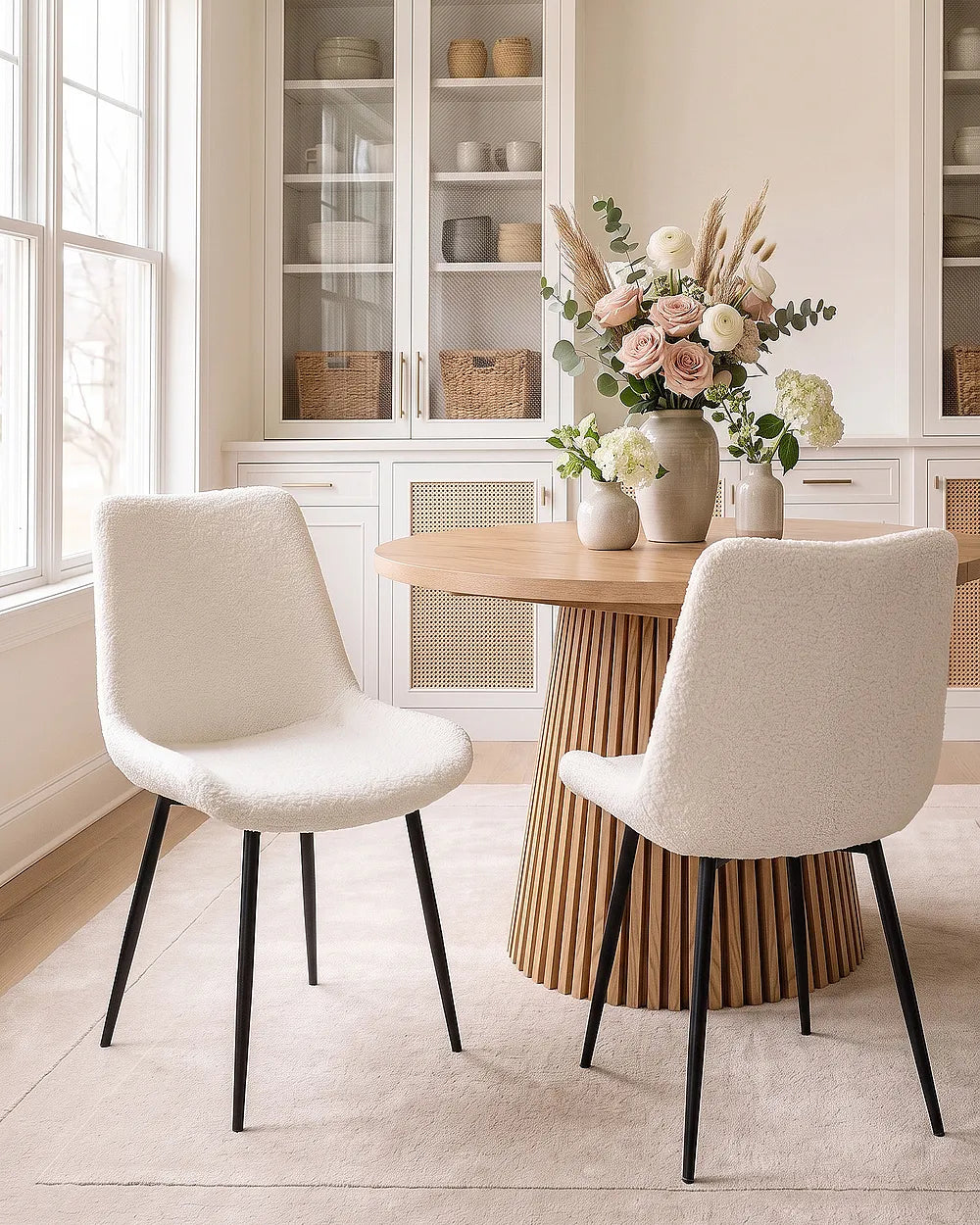 Two modern upholstered dining chairs with textured cream bouclé seats and black tapered legs, positioned around a round wooden table in a bright, neutral-toned dining room.