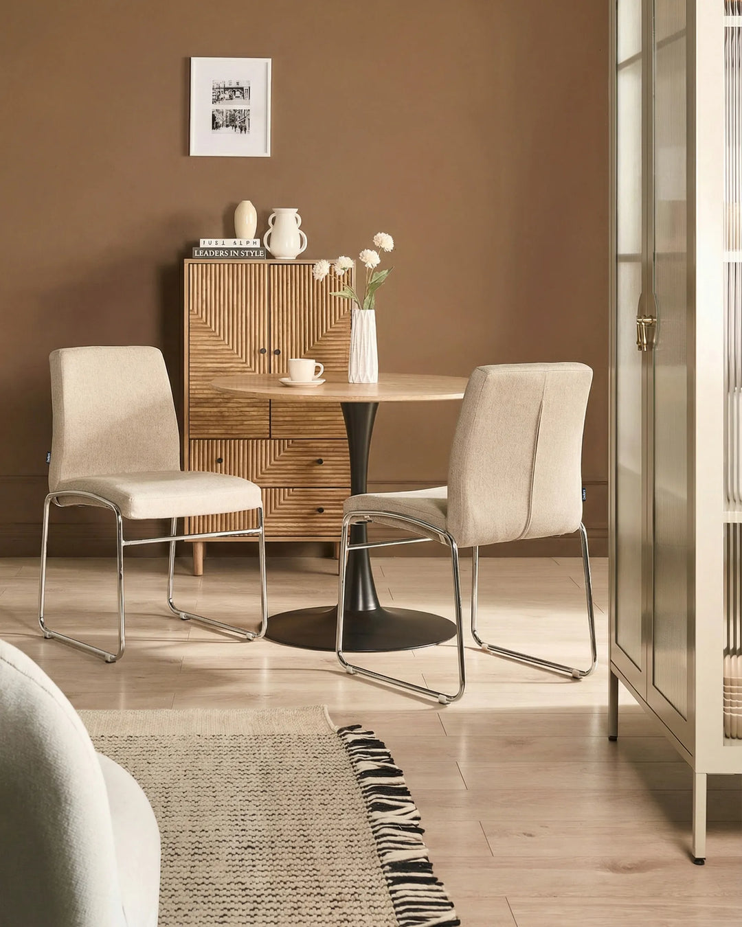 Dining area with beige chairs, wooden cabinet, and a vase with flowers on a round table.