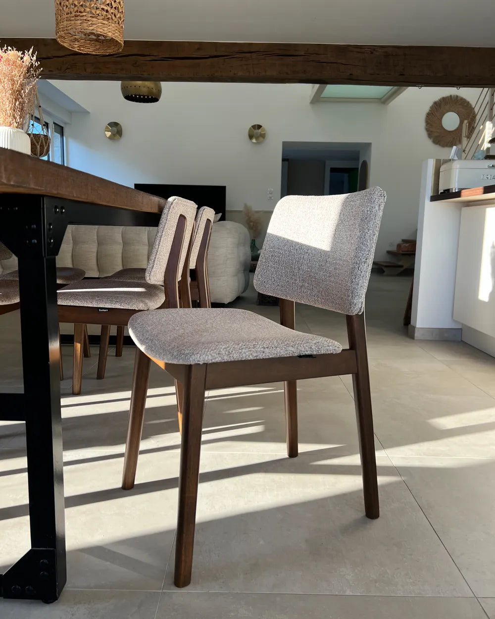 A modern dining chair with a textured light gray upholstered seat and backrest, supported by a dark brown wooden frame, positioned in a bright, contemporary dining room.