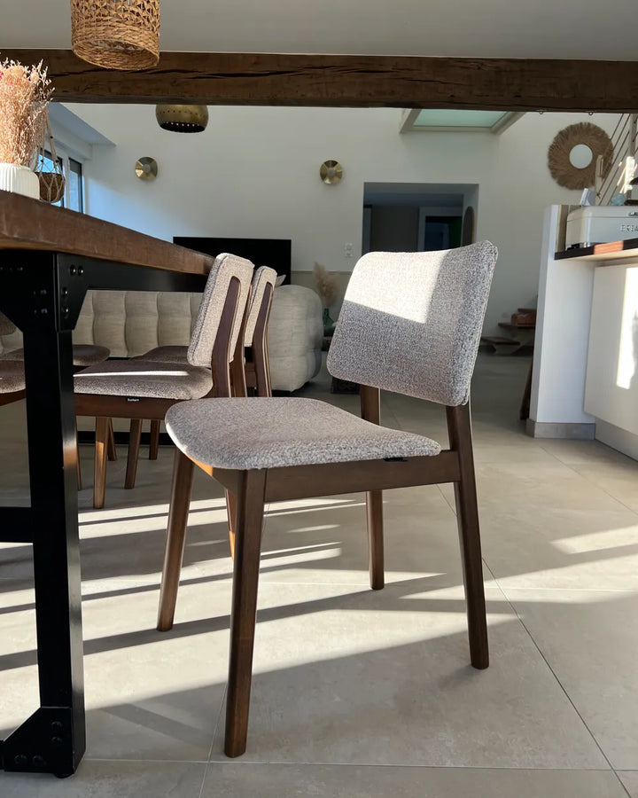A modern dining chair with a textured light gray upholstered seat and backrest, supported by a dark brown wooden frame, positioned in a bright, contemporary dining room.