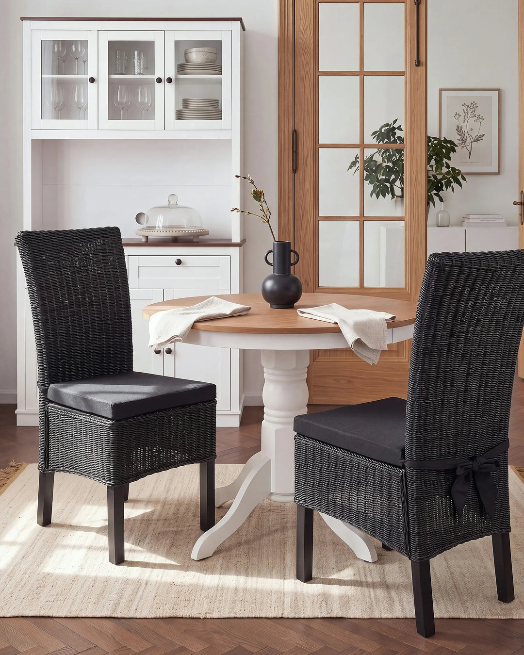 A dining set featuring a round wooden table with a white pedestal base and two black woven wicker chairs with black seat cushions, styled in a bright dining room setting.