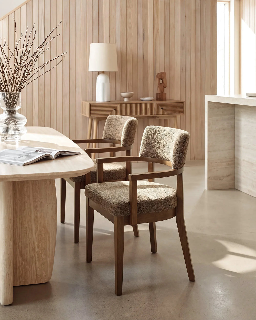 Two upholstered dining chairs with dark wood frames and textured beige fabric, shown in a warm, modern dining room setting with wood-paneled walls and a light wood table.