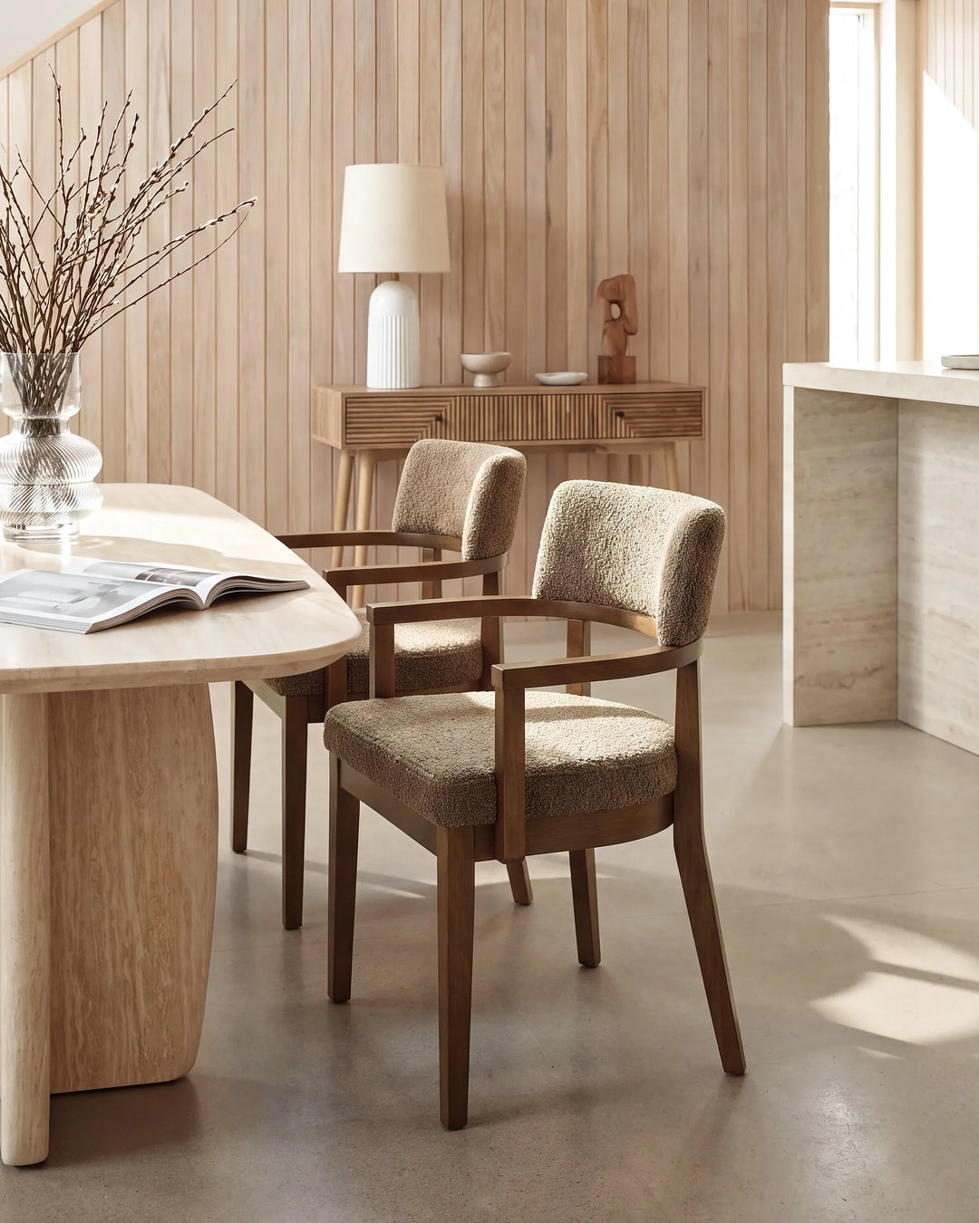 Two upholstered dining chairs with dark wood frames and textured beige fabric, shown in a warm, modern dining room setting with wood-paneled walls and a light wood table.