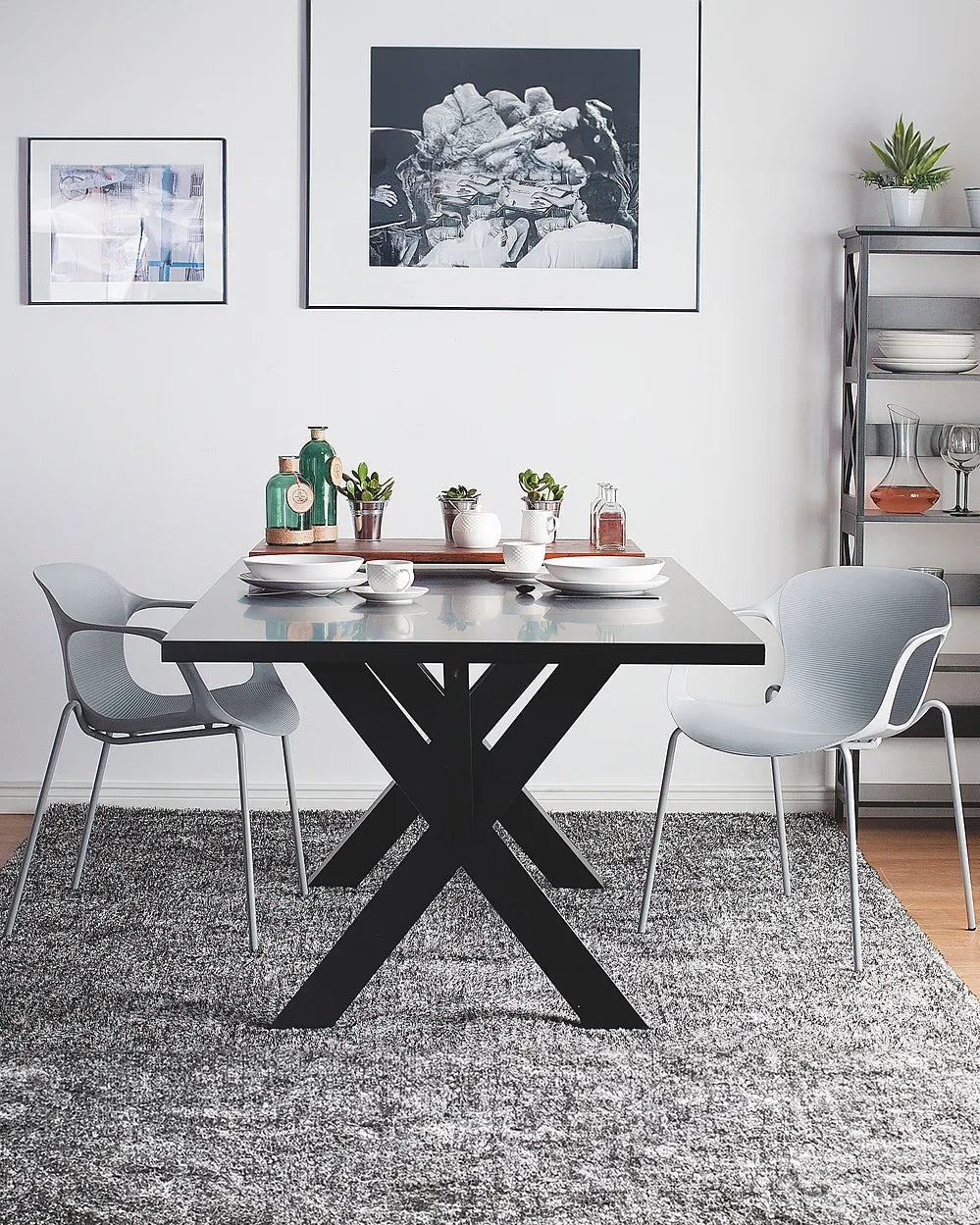 A modern dining room setup featuring a rectangular black table with a bold X-shaped base, flanked by two light gray chairs. The scene is styled with white plates, small potted plants, and decorative bottles on the tabletop, set on a textured gray rug against a white wall with framed art.