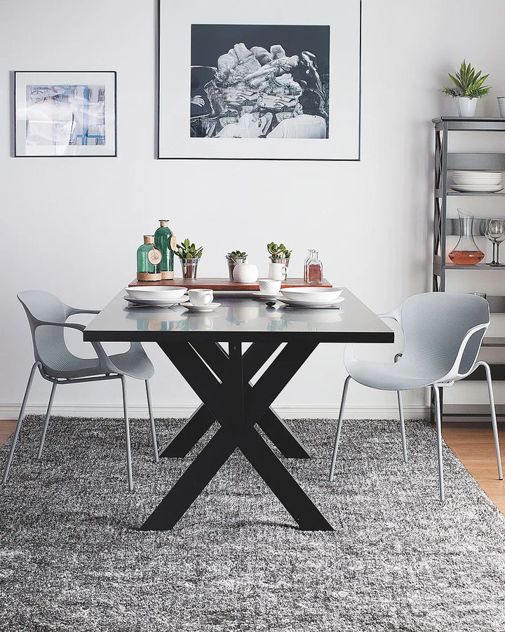 A modern dining room setup featuring a rectangular black table with a bold X-shaped base, flanked by two light gray chairs. The scene is styled with white plates, small potted plants, and decorative bottles on the tabletop, set on a textured gray rug against a white wall with framed art.