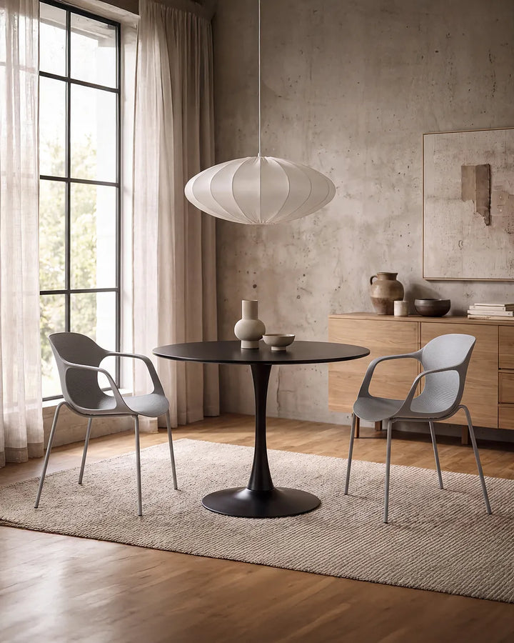 A modern dining room scene featuring a round black pedestal table with two gray curved armchairs, set on a textured beige rug in a room with a concrete wall, wood floors, and a white paper lantern pendant light.
