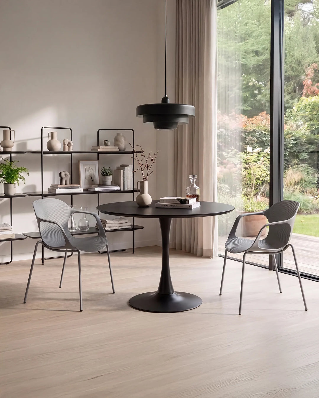A modern dining setup in a bright, minimalist room featuring a round black table with a tulip base, two gray molded chairs, and a black pendant light. Large windows reveal a green garden outside, and a black shelving unit stands against the wall.
