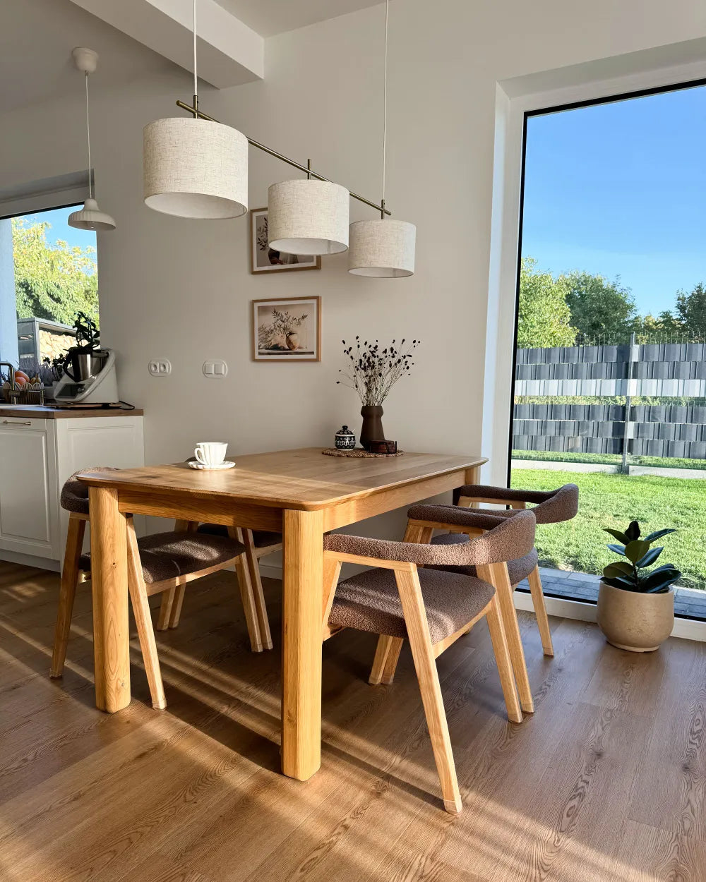 A modern dining room setup featuring a light oak wood rectangular table with four matching wooden chairs that have brown upholstered seats, set in a bright room with a large window overlooking a green lawn.