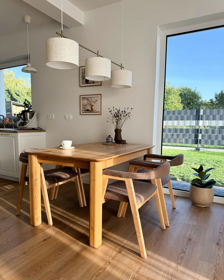 A modern dining room setup featuring a light oak wood rectangular table with four matching wooden chairs that have brown upholstered seats, set in a bright room with a large window overlooking a green lawn.