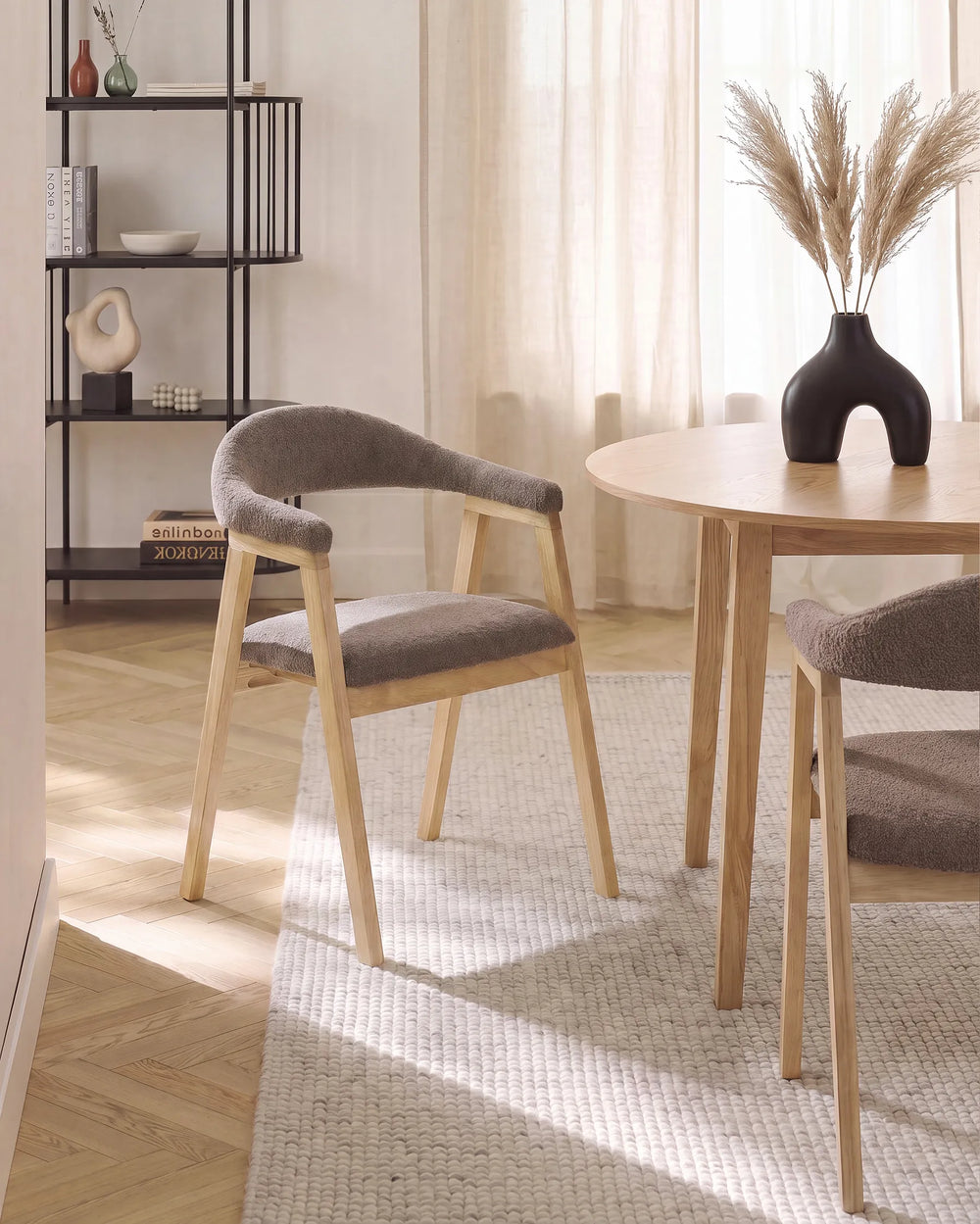 A dining room scene with a round light wood table and two upholstered wooden chairs with gray fabric seats, placed on a textured rug in a bright room with curtains and a black shelving unit in the background.