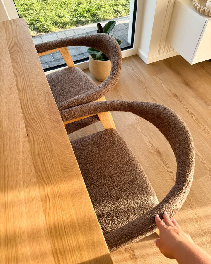 Two modern dining chairs with textured brown bouclé upholstery and curved wooden frames, shown next to a light oak wood table in a bright room with a window and wood floor.