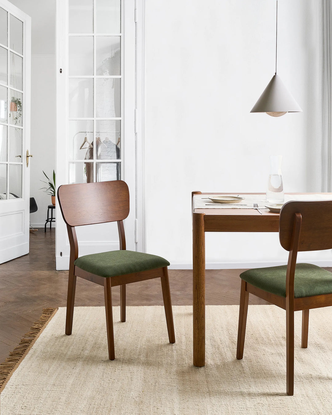 A dining room scene with two wooden chairs featuring green upholstered seats, arranged around a wooden table on a light beige rug in a bright, modern interior.