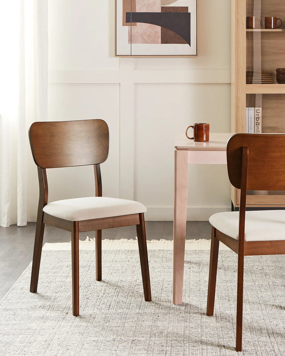 Two mid-century modern wooden dining chairs with cream upholstered seats, shown in a styled dining room setting next to a light pink table.