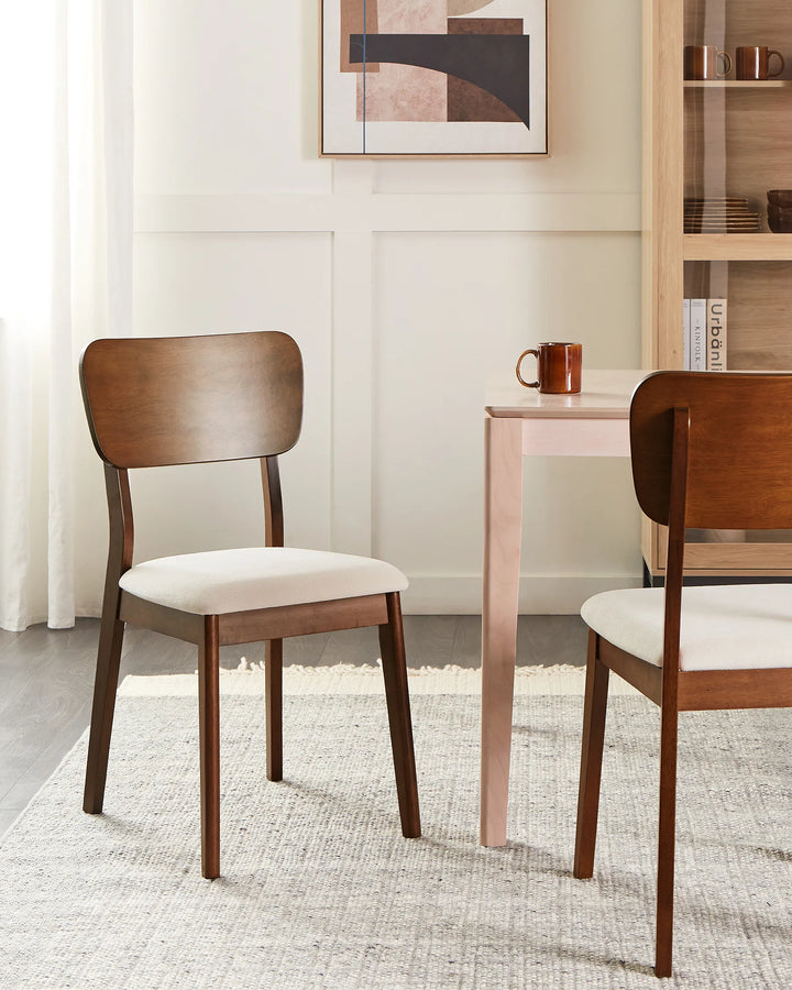 Two mid-century modern wooden dining chairs with cream upholstered seats, shown in a styled dining room setting next to a light pink table.