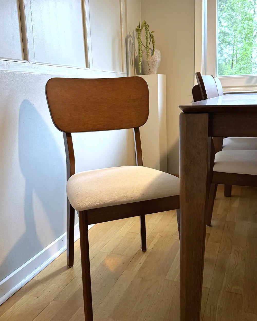 Wooden chair with white cushion next to a wooden dining table in a bright room with large windows.