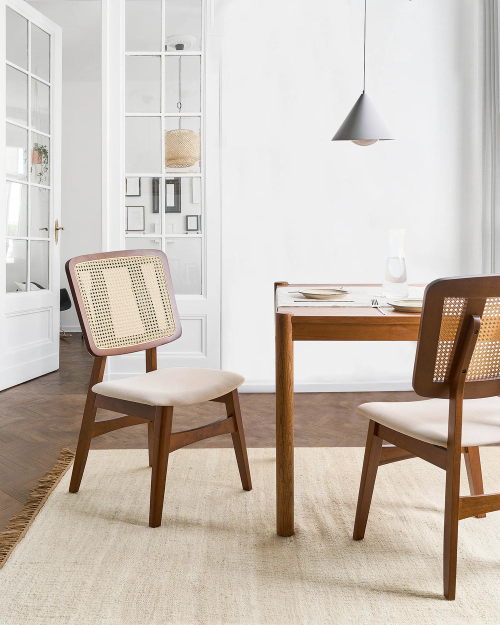 A dining room scene featuring two mid-century modern wooden dining chairs with woven cane backs and light beige upholstered seats, positioned around a wooden dining table on a textured cream rug.