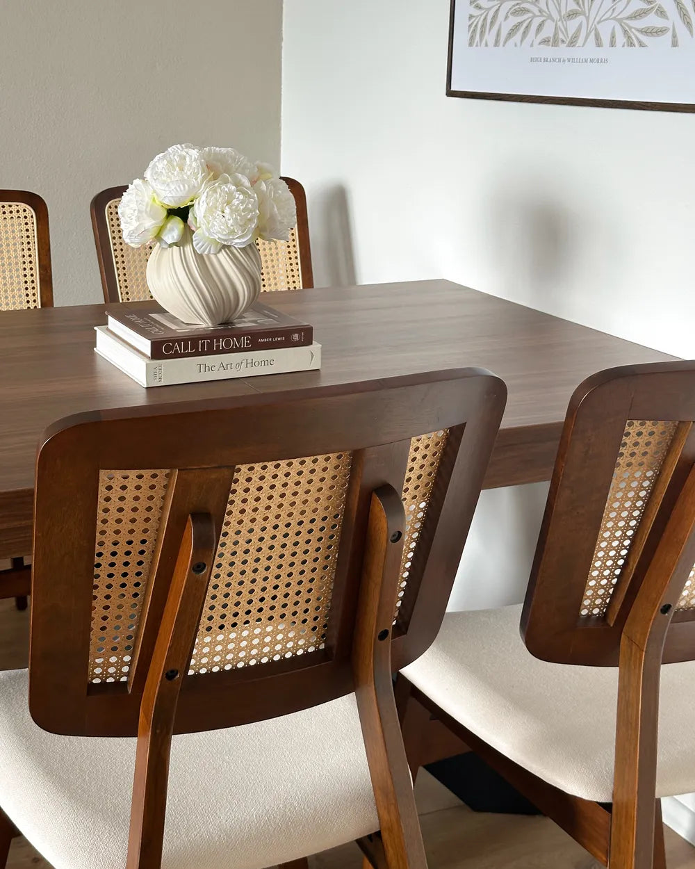 Dining table with books and a vase of flowers, surrounded by wooden chairs with wicker backrests.